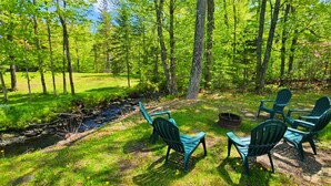 Un terrain de camping avec des chaises Adirondack vertes disposées autour d’un foyer, le tout entouré d’une pelouse et d’un petit ruisseau.