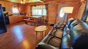 L’intérieur chaleureux d’un chalet avec un plancher en bois, un canapé en cuir noir, une table à manger et un coin cuisine doté de meubles en bois et d’un réfrigérateur noir.