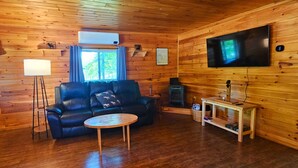 L’intérieur chaleureux d’un chalet avec un canapé en cuir, une table basse ronde, un téléviseur à écran plat et un poêle à bois.
