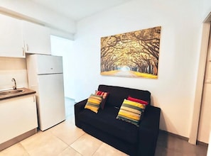 A compact living space with a kitchenette, a black sofa with colorful cushions, and a painting of a tree-lined pathway on the wall.