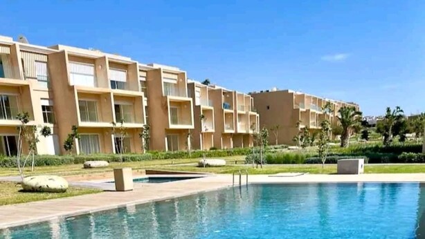 Une piscine à l’eau d’un bleu cristallin, entourée d’une terrasse carrelée. Au fond, on aperçoit des immeubles à plusieurs étages dotés de balcons et une pelouse bien entretenue.