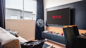 A modern living room with a large flat-screen TV displaying the Netflix logo, a black sofa, a wooden entertainment unit, and a window with curtains. - Interlagos Autodrome Apartment (Vila da Paz São Paulo)