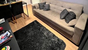 A modern living room with a beige sofa, black rug, and a wall-mounted TV displaying sports content. - Interlagos Autodrome Apartment (Vila da Paz São Paulo)