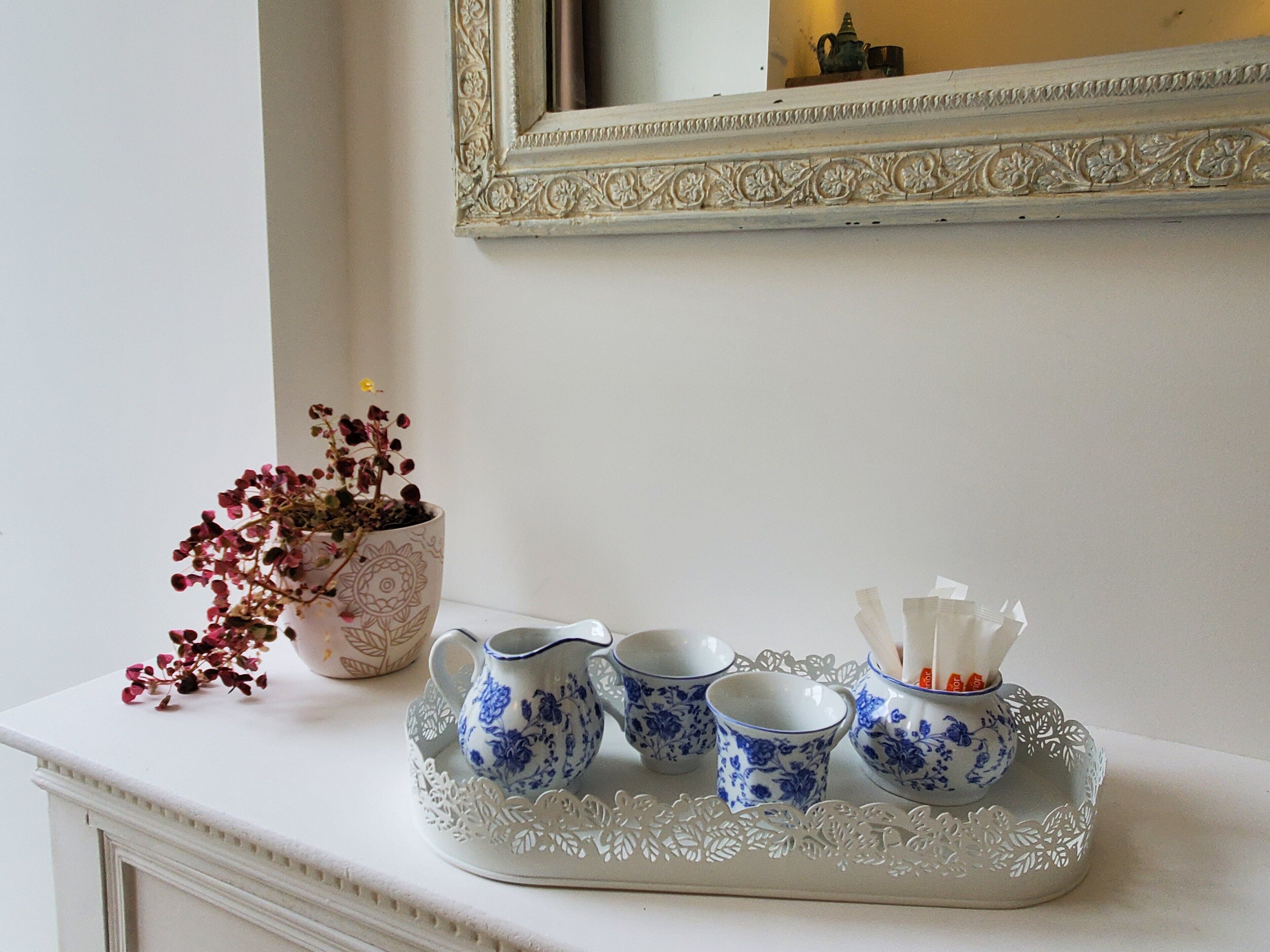 Une table blanche sur laquelle repose un plateau décoratif contenant trois tasses à motifs floraux bleues et blanches et une carafe, ainsi qu’une plante en pot aux fleurs rouges.