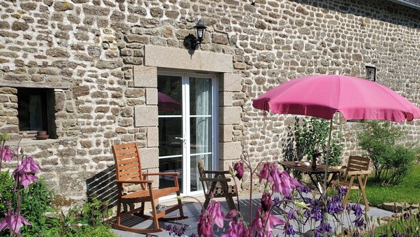 Un bâtiment en pierre avec une chaise berçante en bois et une table surmontée d’un parasol rose.