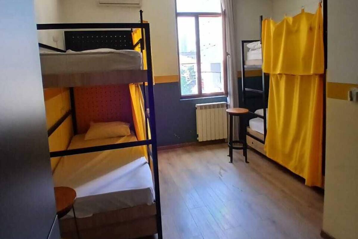 A dormitory room with bunk beds, a small table, a window with curtains, and a radiator.
