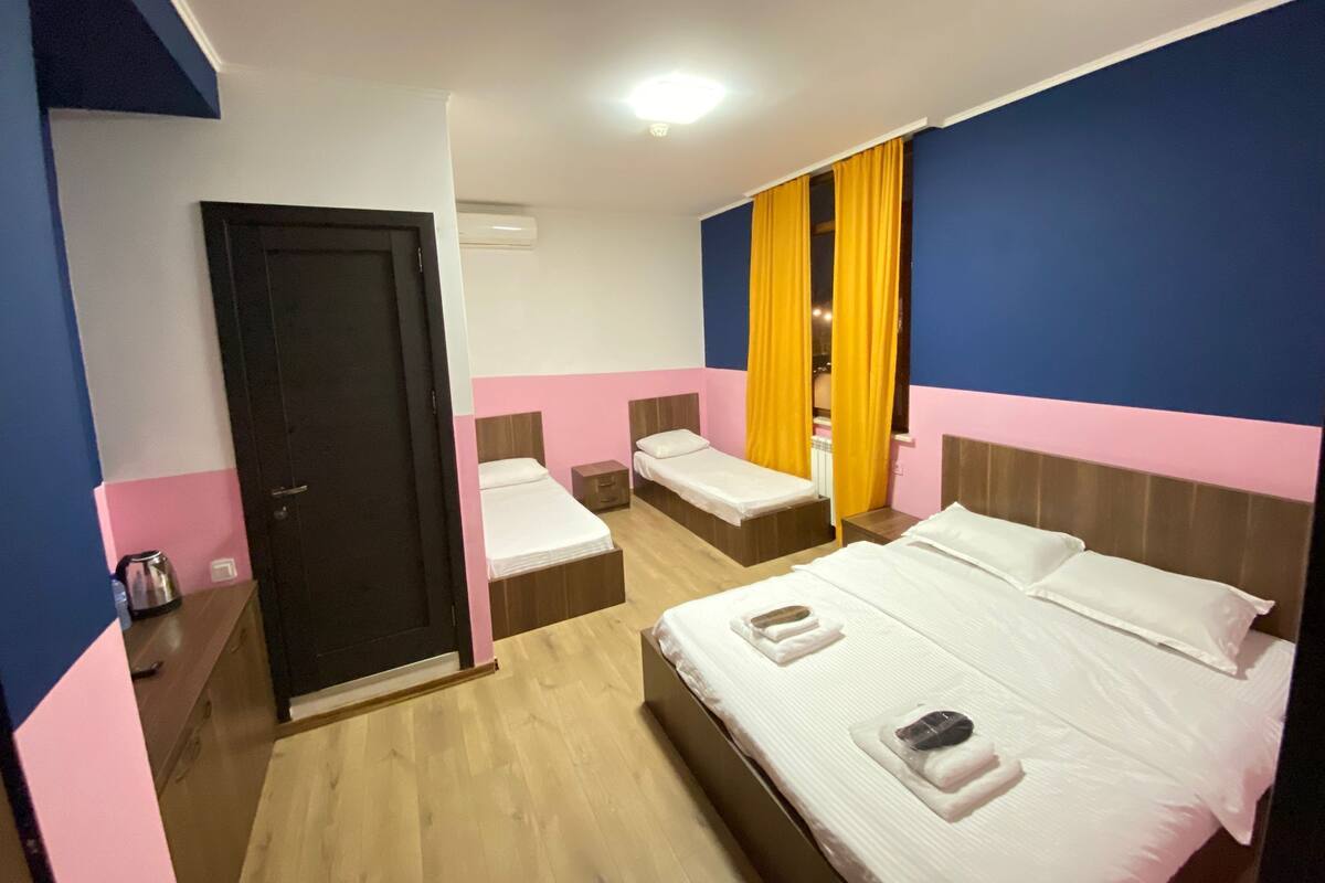 A hotel room with two single beds, a wooden floor, a black door, and a window with yellow curtains.