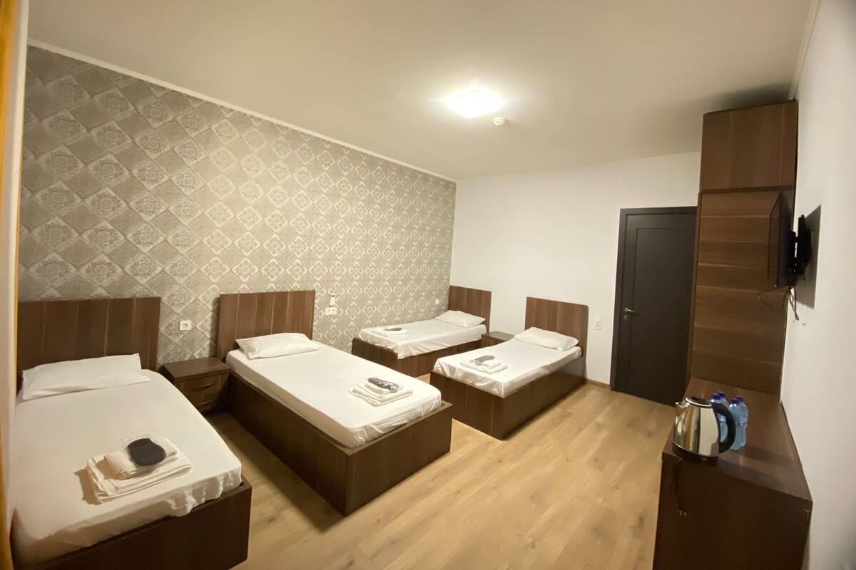 A hotel room with three single beds, a wooden wardrobe, a flat-screen TV mounted on the wall, and a patterned wallpaper on the left wall.