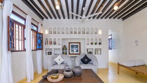 Una habitación con techo de madera, dos sofás grises con cojines, una mesa de madera con cerámica y una cama con colcha estampada.