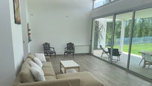 A spacious living room with a beige sofa, two armchairs, a coffee table, and a sliding glass door leading to an outdoor area with a lawn and chairs. - House in Meadows of San Lorenzo - San Lorenzo Chico / Salta (Salta)