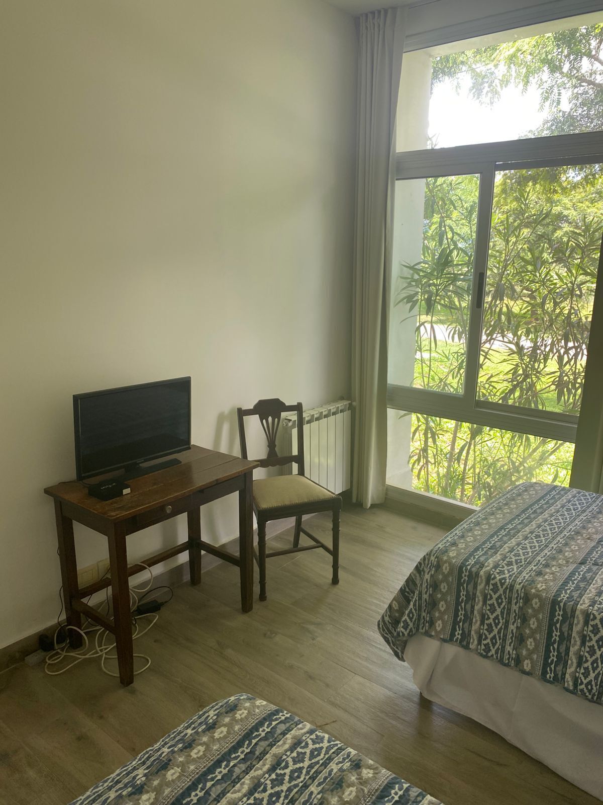 A hotel room with a TV on a wooden table, a chair, two beds with patterned bedding, and a large window with a view of greenery.