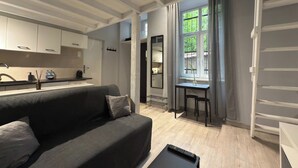 Ein modernes Wohnzimmer mit einer grauen Couch, einem kleinen Tisch und einer Küchenzeile mit weißen Schränken und einem Fenster mit Vorhängen.