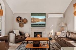 A living room with a fireplace, a television mounted above it, and a large window with blinds.
