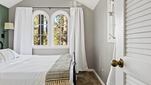 Une chambre à coucher avec un grand lit, deux fenêtres avec des rideaux, une lampe et une porte dotée d’une poignée en laiton.