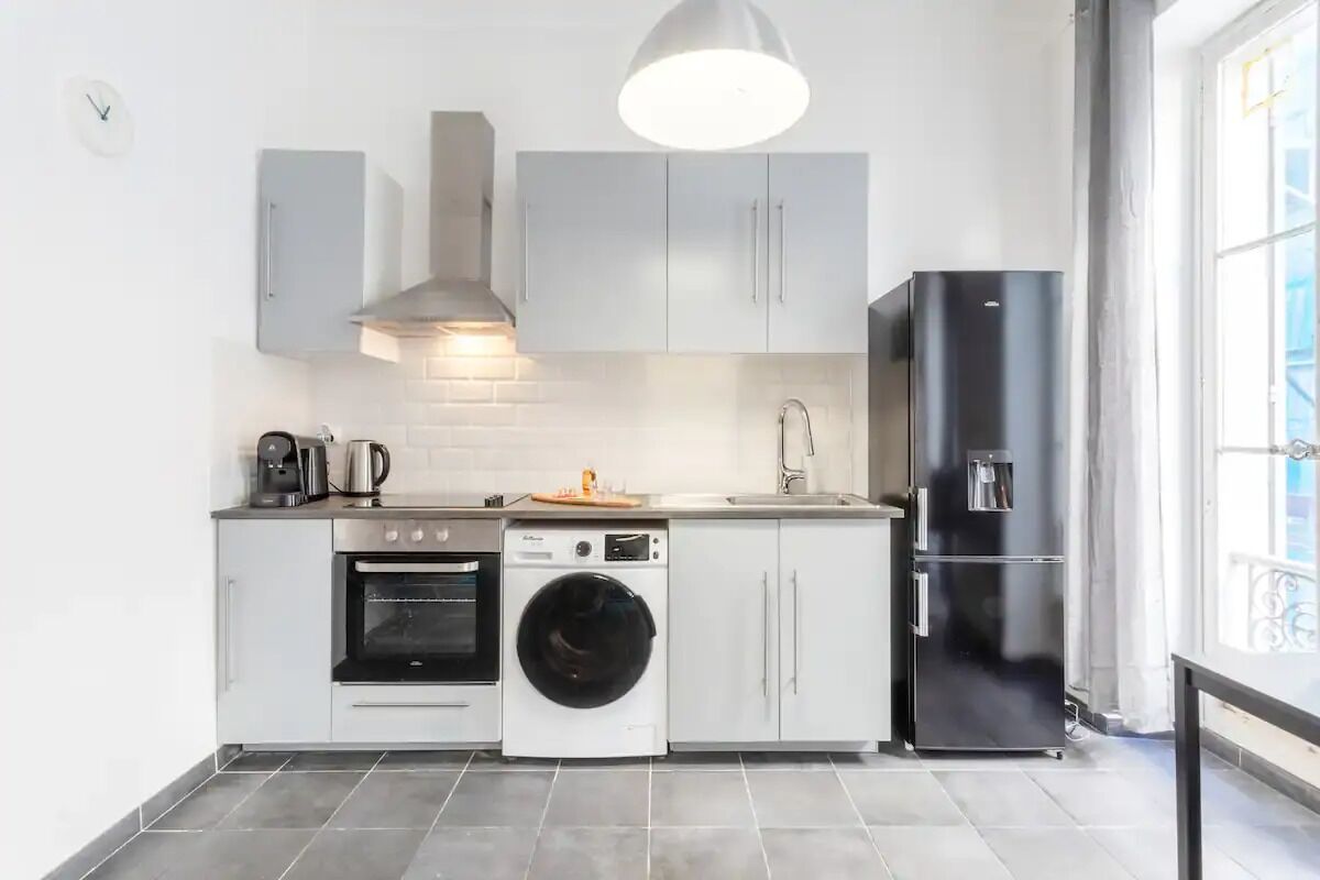 Une cuisine compacte avec des meubles blancs, un réfrigérateur noir, un lave-linge et une machine à café.