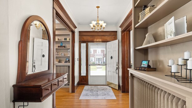 Un couloir avec un plancher en bois, une porte blanche, un lustre et des étagères remplies de livres et d’objets décoratifs.
