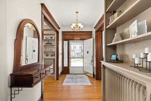 Un couloir avec un plancher en bois, une porte blanche, un lustre et des étagères remplies de livres et d’objets décoratifs.