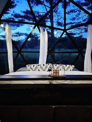 A bed with a patterned pillow, a small sign reading 'HOME,' and a view of trees through a geometric window structure.