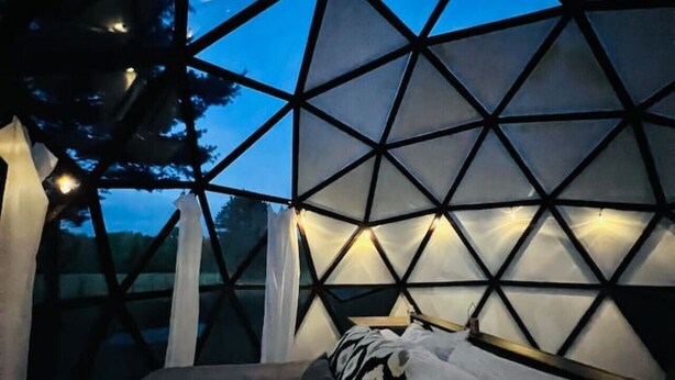 A room with a geometric ceiling structure, a bed with a patterned blanket, and a view of the night sky.