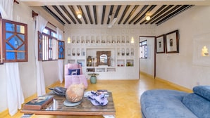 A room with a wooden ceiling, a blue sofa, a glass coffee table, and a wall with decorative shelves. - Pate Suite (Shela)