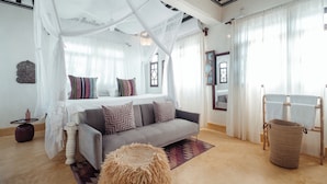 A bedroom with a canopy bed, a grey sofa, a woven ottoman, and a wooden laundry stand. - Pate Suite (Shela)
