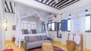A bedroom with a bed, a grey sofa, a woven stool, and a wooden drying rack. - Pate Suite (Shela)
