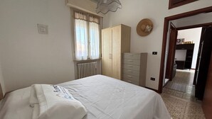 A bedroom with a bed, a wardrobe, a radiator, a window with curtains, and a door leading to another room.