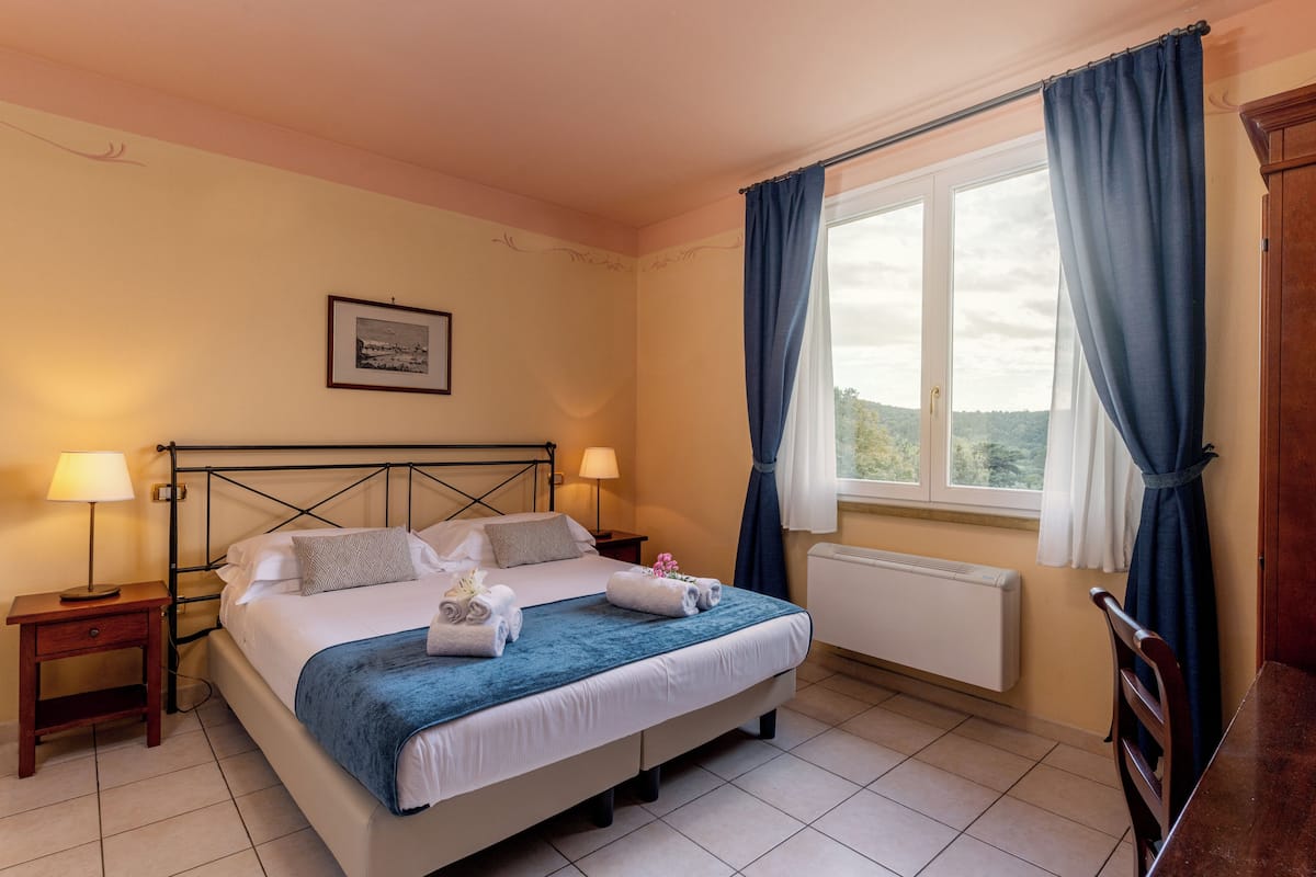 Un letto rifatto con cuscini e una coperta blu, un comodino con una lampada, e una finestra con tende.