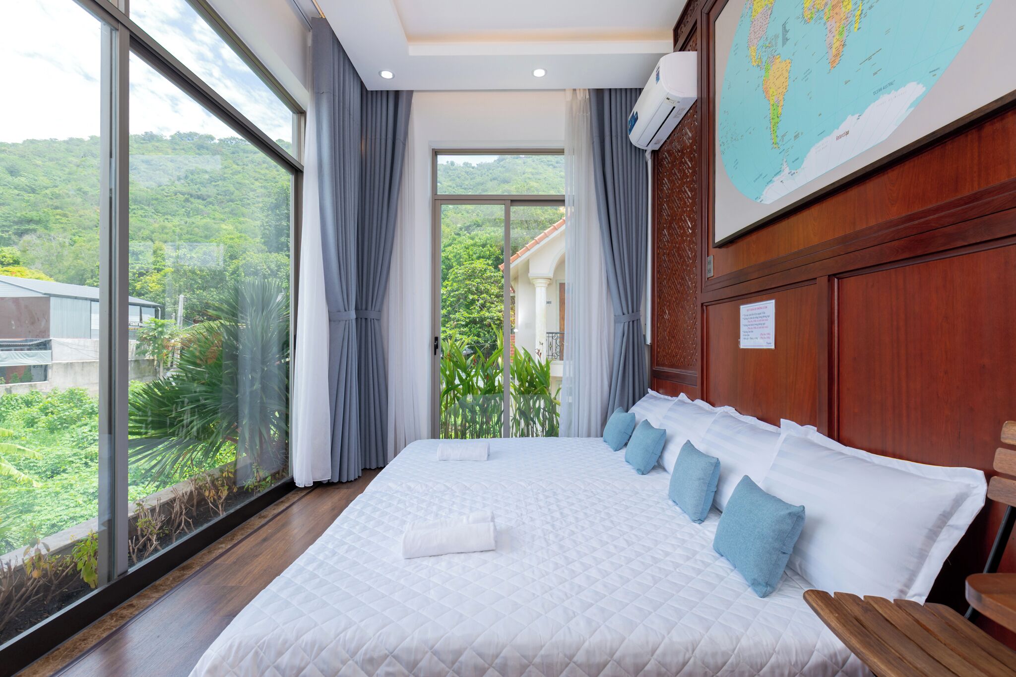 A hotel room with a large bed, a view of greenery outside, and a world map on the wall.