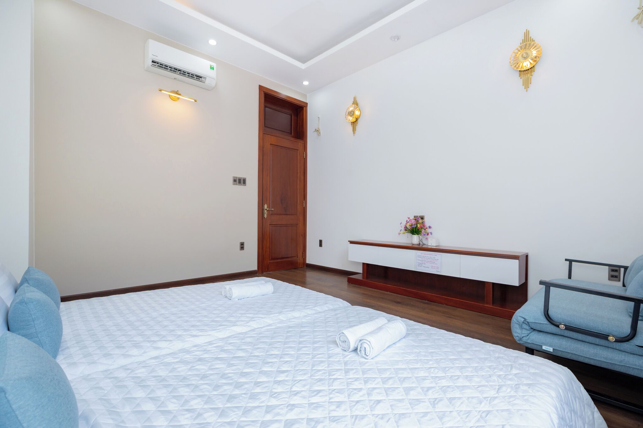 A hotel room with a large bed, a bedside table, a chair, and an air conditioning unit.