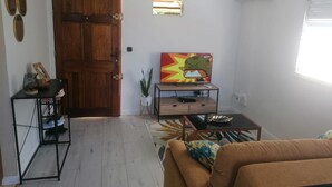 A living room with a wooden door, a TV on a wooden stand, a sofa, a coffee table, and a small table with a basket.