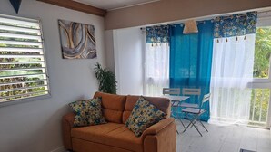 A living room with a brown sofa, floral cushions, a small round table, and a patterned rug. There is a window with blinds, a plant, and a blue curtain.