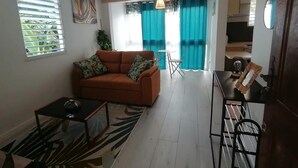 A living room with a sofa, coffee table, and a small table with a lamp. There is a view of a kitchen and a dining area with a table and chairs.