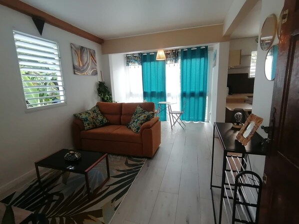 A living room with a sofa, coffee table, and a small table with a lamp. There is a view of a kitchen and a dining area with a table and chairs.