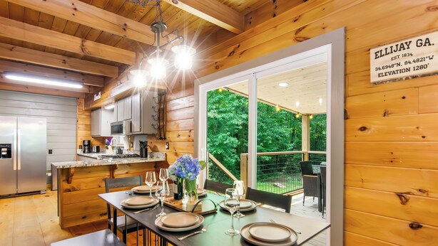Interior de una cabaña de madera con comedor para cuatro, cocina equipada con electrodomésticos modernos y un amplio ventanal con vistas al bosque.