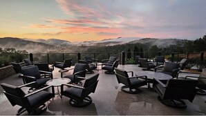 Un patio al aire libre con mesas y sillas redondas, con vista a un paisaje montañoso al atardecer.
