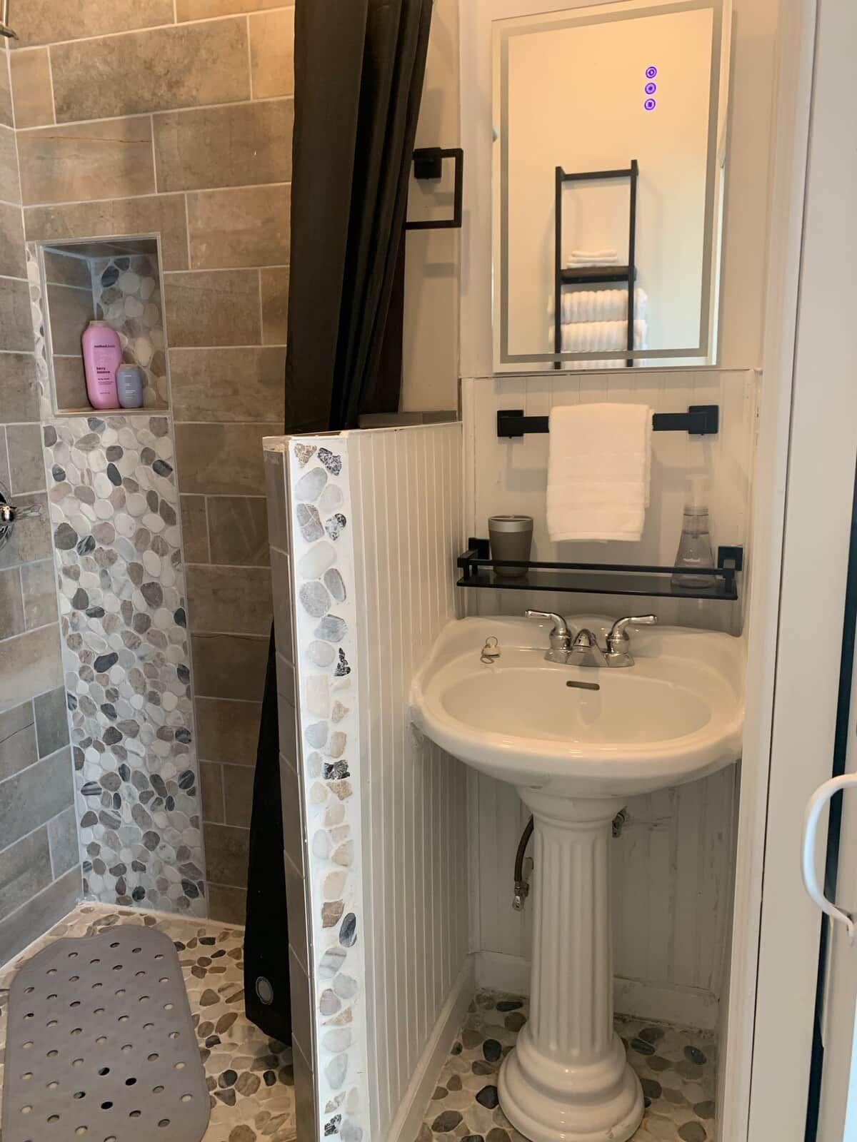 A bathroom with a white pedestal sink, a mirror above it, and a shower area with stone tiles.