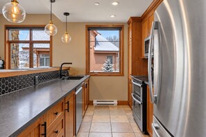 A modern kitchen with a stainless steel refrigerator, wooden cabinets, a black countertop, and a window with a view of a snowy landscape. - Tremblant Prestige - Altitude 170-9 (Mont-Tremblant)