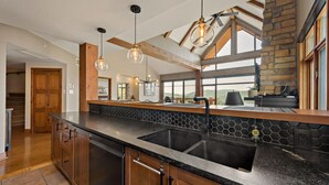 A modern kitchen with a large island, black countertops, wooden cabinets, a stone backsplash, and a view of the outdoors through large windows. - Tremblant Prestige - Altitude 170-9 (Mont-Tremblant)