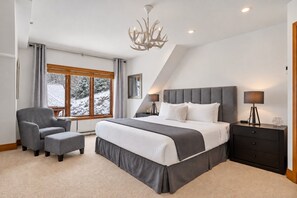 Une chambre moderne avec un grand lit, un fauteuil à revêtement gris, une table de chevet avec une lampe, et une vue sur un paysage enneigé par une grande fenêtre.