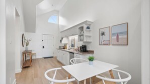 Una cocina luminosa con armarios blancos, un comedor con mesa y sillas blancas, y una pequeña mesita con una planta.