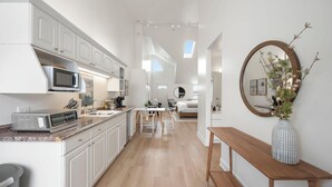 Una cocina moderna con muebles blancos, un microondas y una cafetera. Hay un comedor con una mesa y sillas de madera. Un espejo redondo cuelga en la pared, y un florero con flores está sobre una consola de madera.