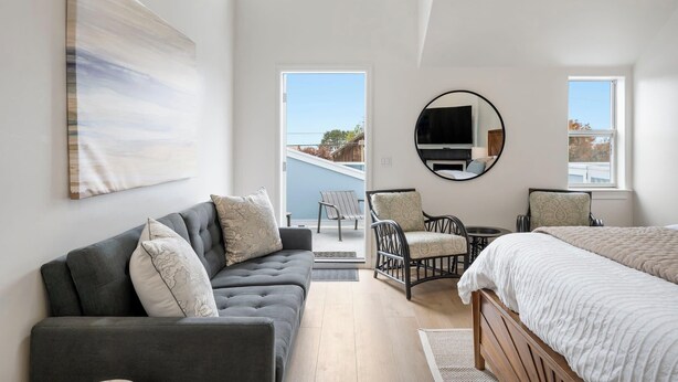 Una habitación de hotel moderna con un sofá gris, dos sillones, una cama, un espejo redondo y un cuadro grande en la pared.