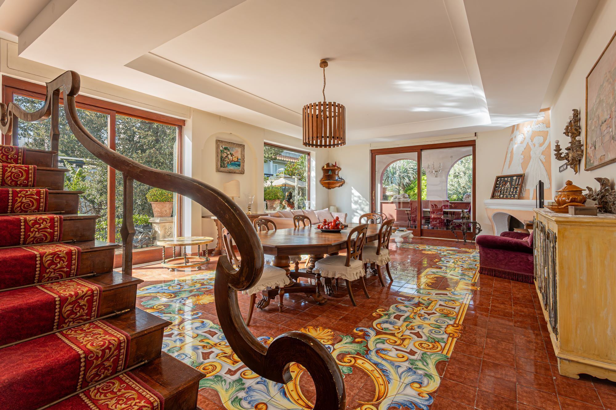 Ampla sala de estar com uma escadaria de madeira grande, uma mesa de jantar e um chão colorido com azulejos.