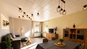 A modern living room with a sofa, a glass coffee table, a dining table with chairs, and a TV on a wooden cabinet. - Dessau UNESCO vacation apartment "Franz" - for nature and culture lovers (Dessau-Roßlau)