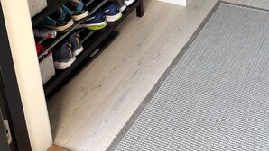 A hallway with a black shoe rack holding various pairs of shoes, a coat hanging on the wall, and a grey rug on the floor.