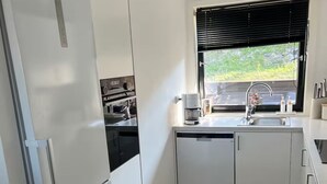 A compact kitchen with white cabinetry, a built-in oven, a dishwasher, and a window with blinds.