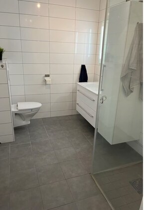 Un baño con azulejos blancos, inodoro, lavabo y zona de ducha.