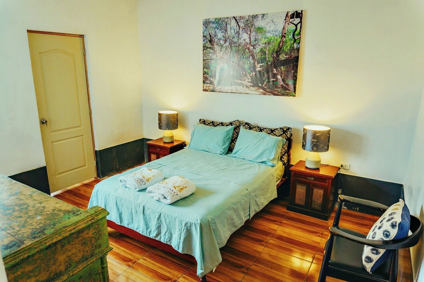 Un dormitorio con una cama, dos mesitas de noche con lámparas, suelo de madera, una silla verde y una puerta.