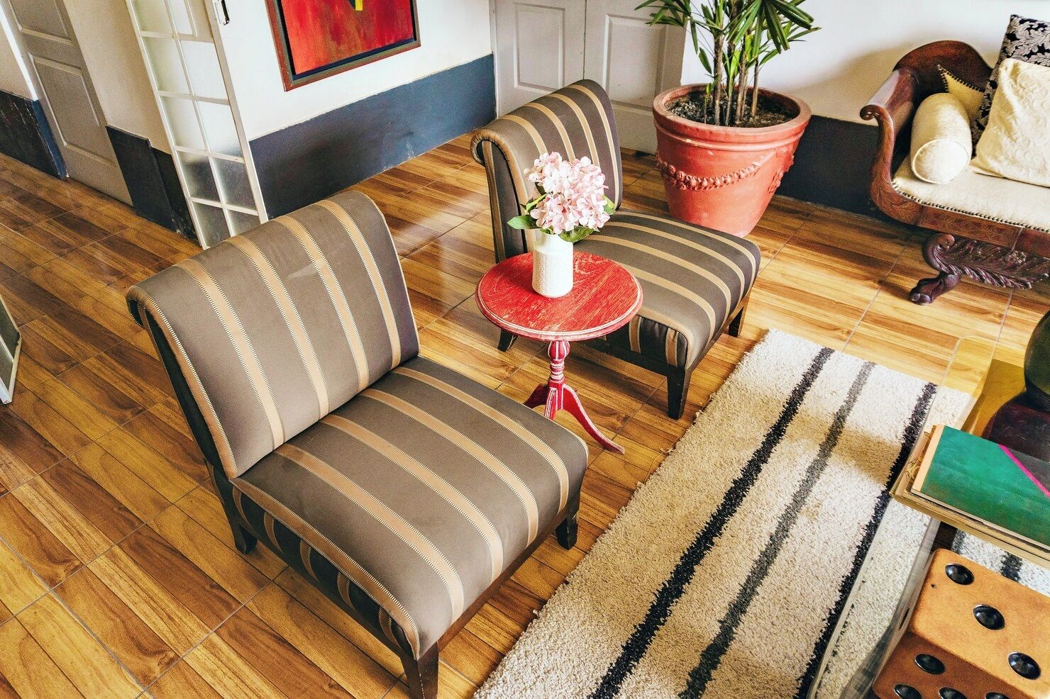A cozy living room with striped sofas, a small red table with a vase of flowers, and a potted plant.